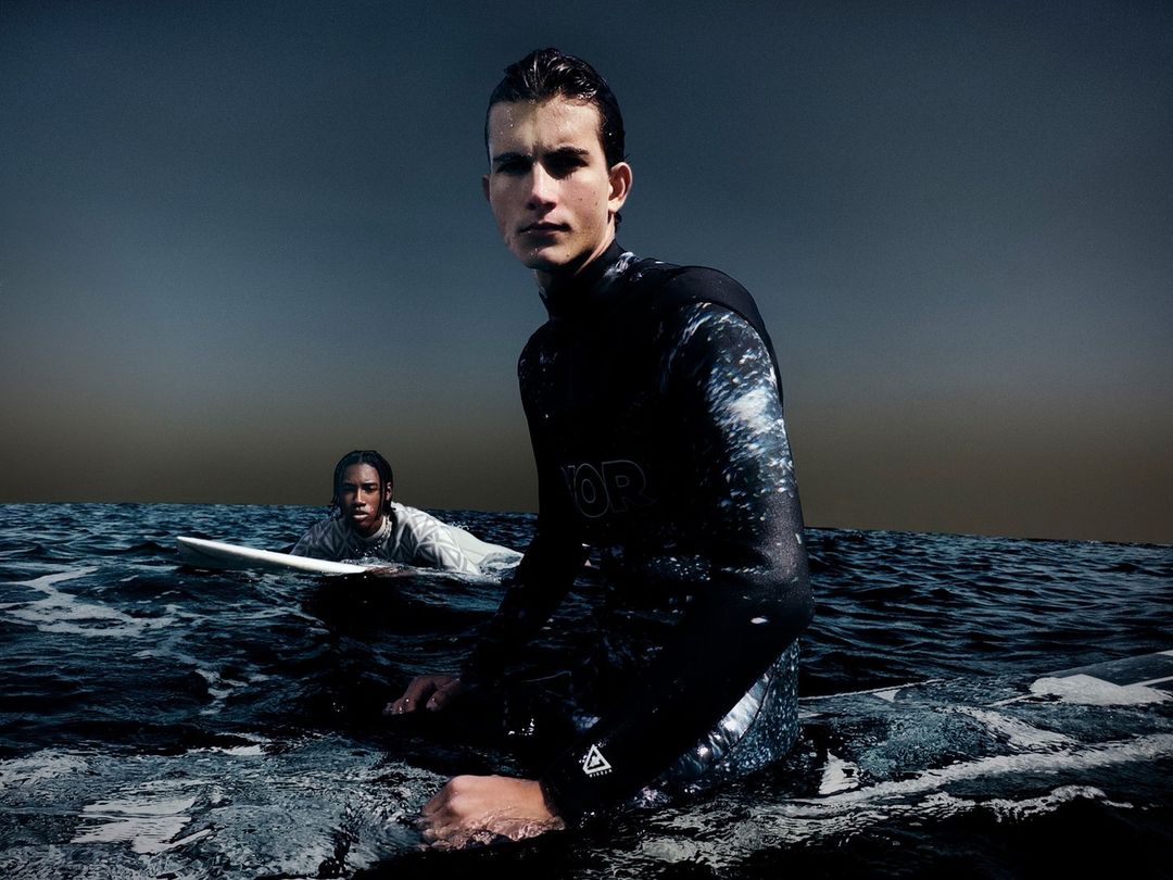 Two surfers wearing wetsuits are in the ocean. One is standing closer to the camera, the other sits on a surfboard further back.