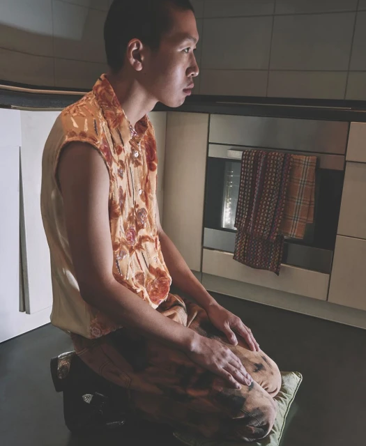 Man sits kneeling indoors wearing orange patterned sleeveless shirt and patterned pants, next to kitchen cabinets and oven.