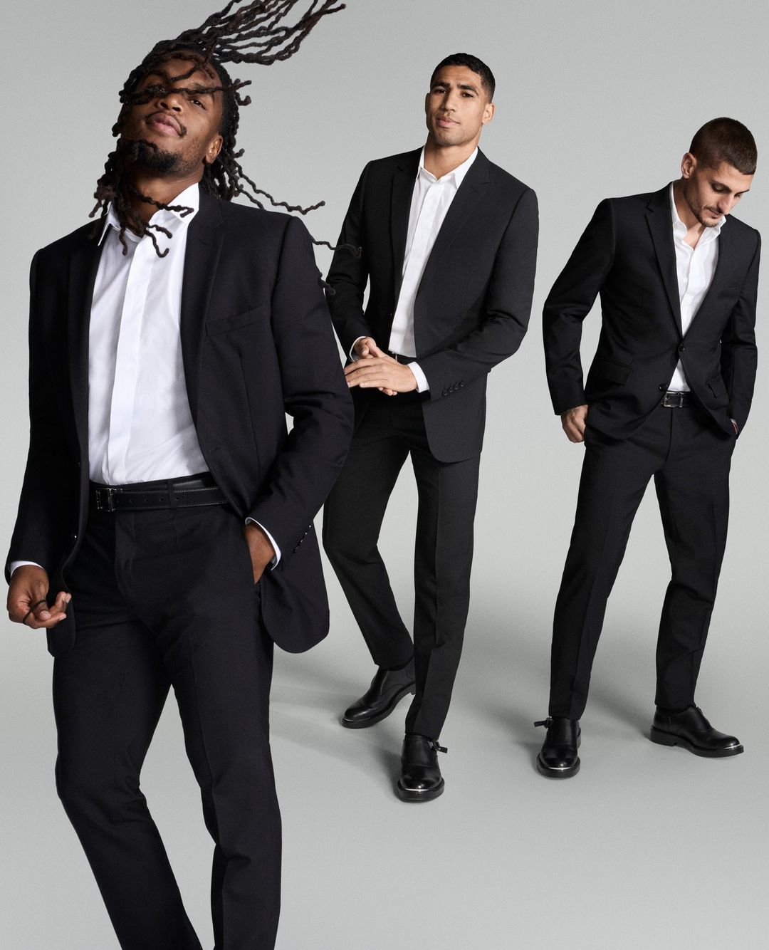 Three adult men in black suits and white shirts pose in a studio setting, showcasing fashion and style with a clean, neutral background.