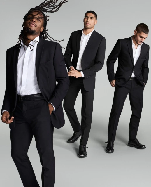 Three adult men in black suits and white shirts pose in a studio setting, showcasing fashion and style with a clean, neutral background.