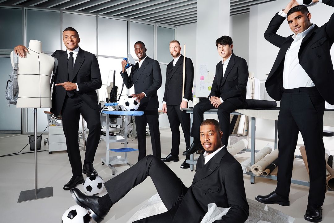 Group of seven stylishly dressed soccer players pose in a bright tailor's studio, surrounded by dress forms, ironing boards, and fabrics.