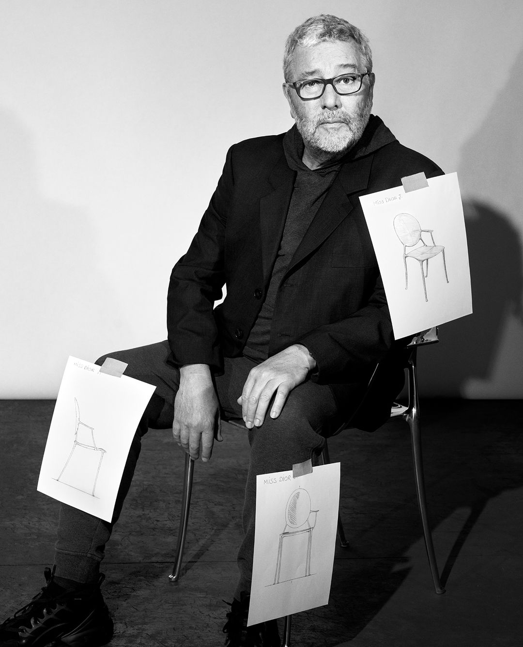Black and white portrait of a seated man with glasses, surrounded by sketches of chairs attached to his clothing and chair.