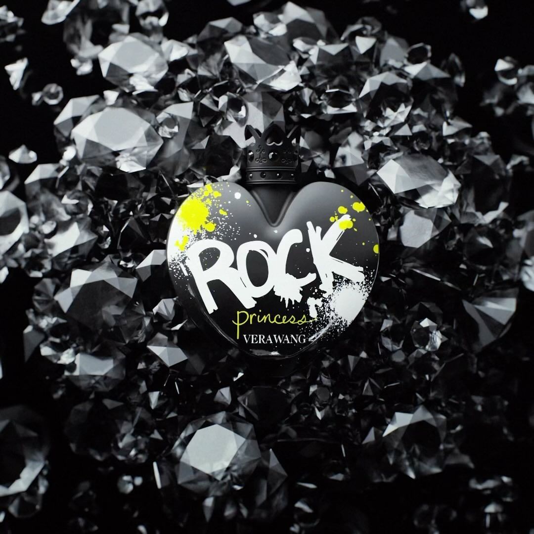 Heart-shaped black perfume bottle of Vera Wang Rock Princess, against a backdrop of faceted black gemstones on a dark surface.