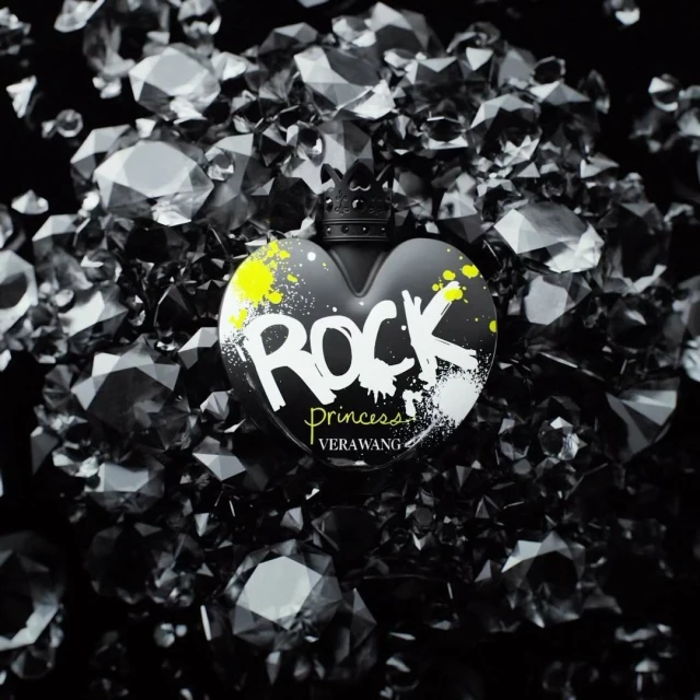 Heart-shaped black perfume bottle of Vera Wang Rock Princess, against a backdrop of faceted black gemstones on a dark surface.