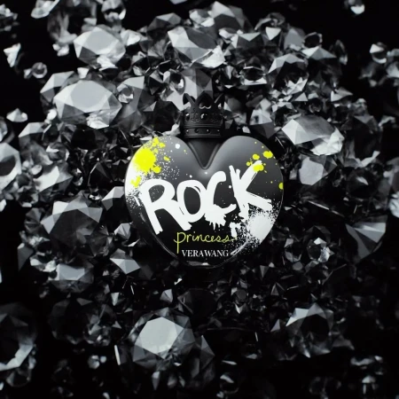 Heart-shaped black perfume bottle of Vera Wang Rock Princess, against a backdrop of faceted black gemstones on a dark surface.