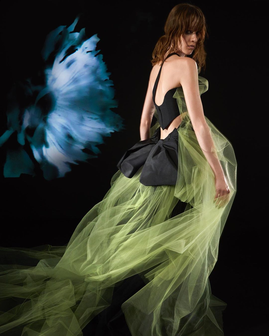 A woman in an evening gown poses in a studio, the gown features a black bodice and a voluminous chartreuse train with floral background.
