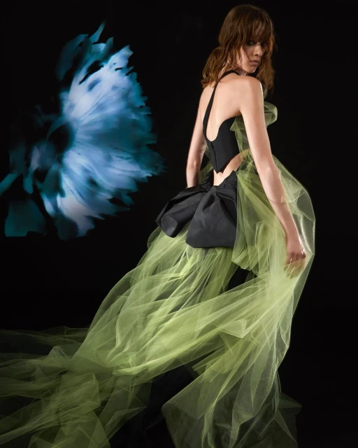 A woman in an evening gown poses in a studio, the gown features a black bodice and a voluminous chartreuse train with floral background.