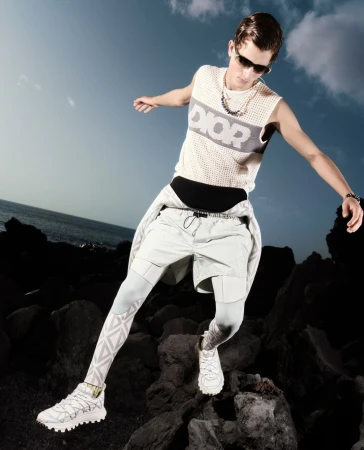 A young male model in sunglasses and a sleeveless mesh Dior top leaps across dark volcanic rocks near the ocean.