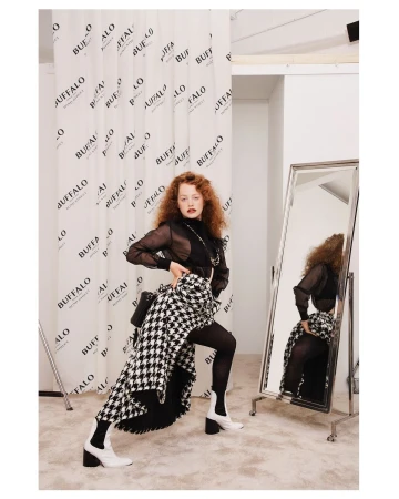 Fashionable woman in sheer top, houndstooth skirt, white boots poses in a studio with a Buffalo logo backdrop and mirror, indoor setting.