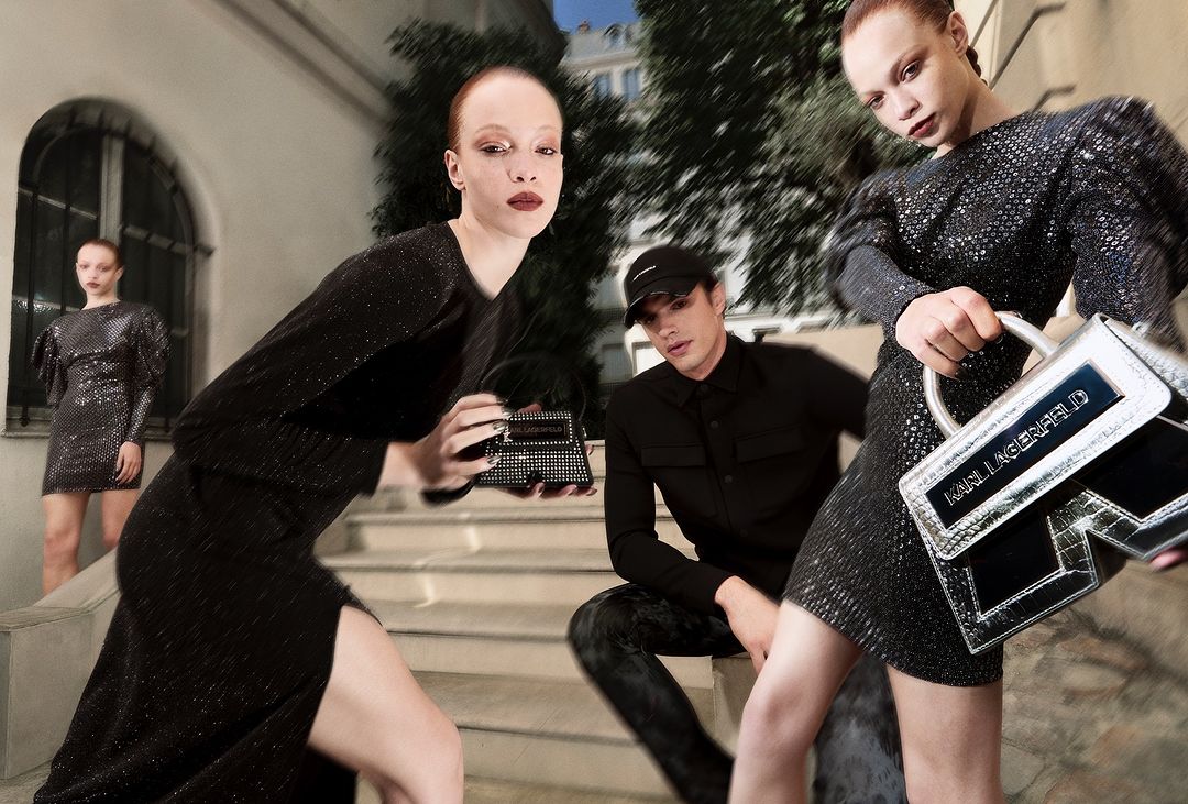 Fashion editorial featuring women in black dresses with a man in black attire, posing on steps in an urban outdoor environment with handbags.