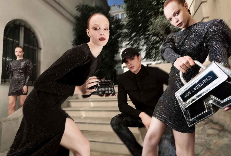 Fashion editorial featuring women in black dresses with a man in black attire, posing on steps in an urban outdoor environment with handbags.