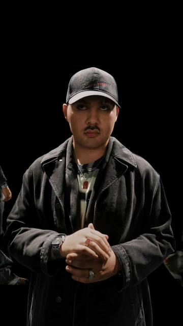 Portrait of an adult male wearing a denim jacket and baseball cap, facing forward against a dark background in an indoor environment.