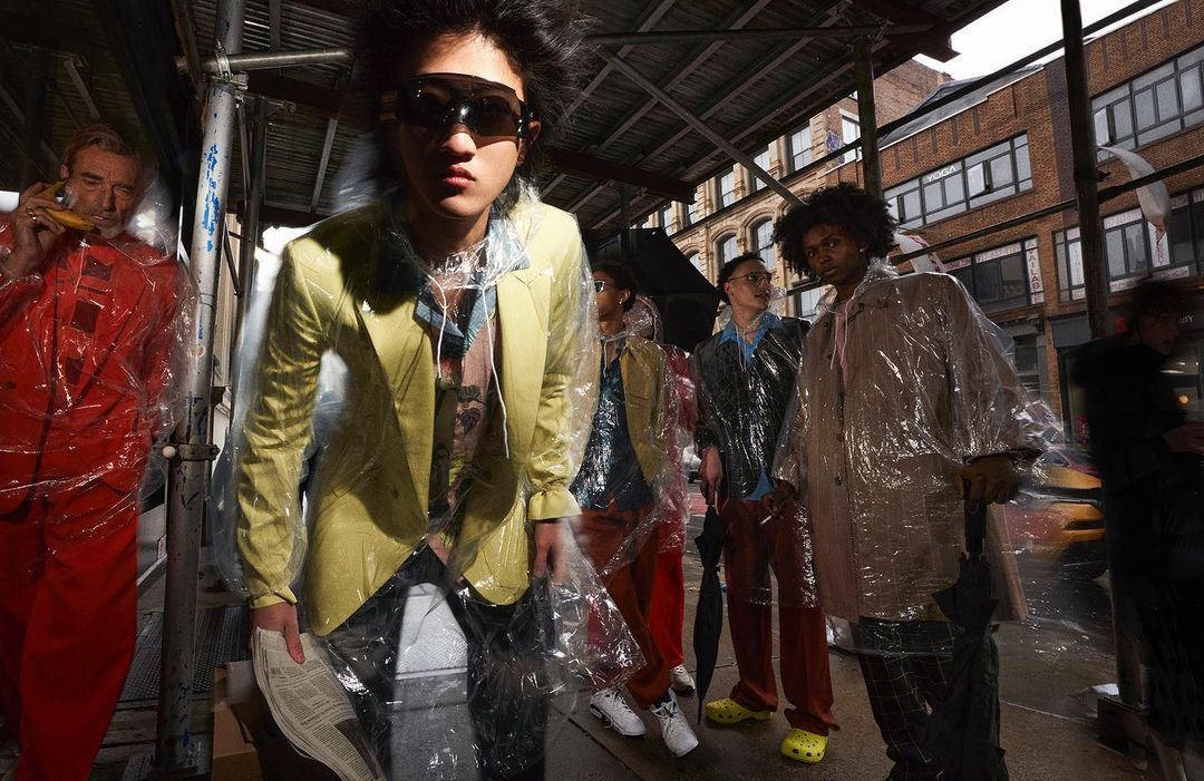 Group of models wearing clear plastic raincoats and diverse casual outfits, posing on a city street with scaffolding and brick buildings.