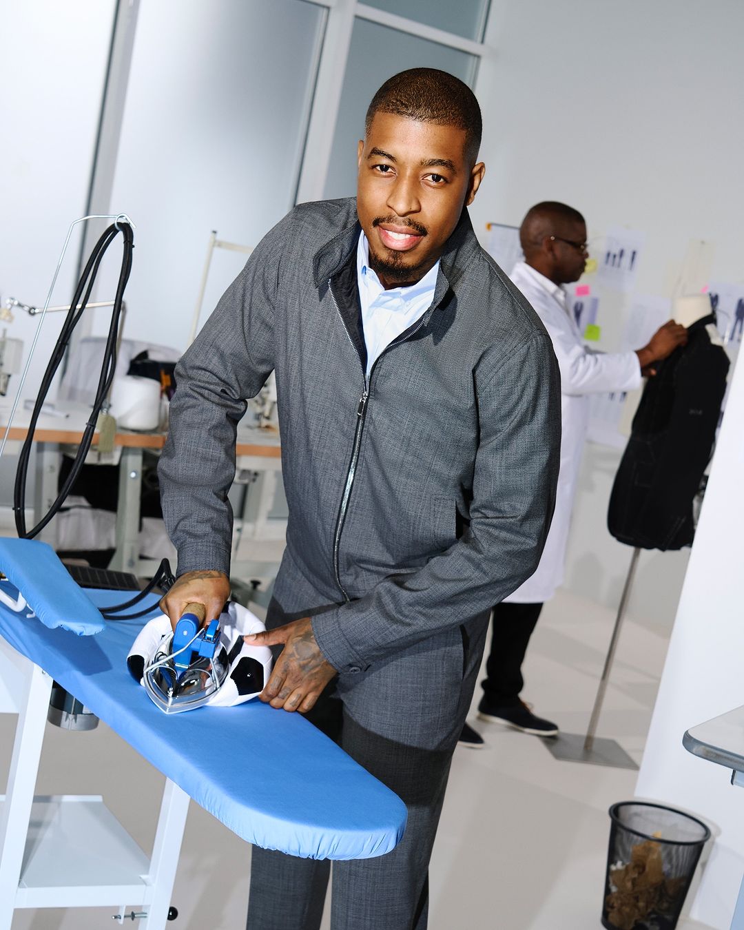 Adult male in a grey suit irons a soccer ball on a blue ironing board indoors in a fashion or design studio with a second man working.