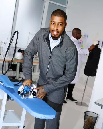 Adult male in a grey suit irons a soccer ball on a blue ironing board indoors in a fashion or design studio with a second man working.