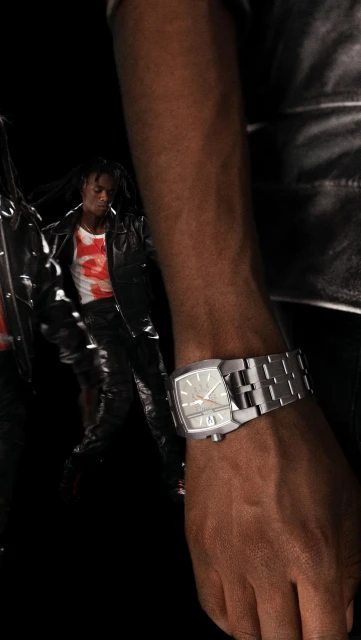 Closeup of a dark-skinned adult wearing a silver watch, with a dark-haired man in the background wearing a black jacket.