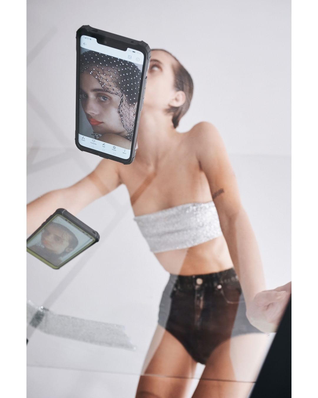 Low angle view of woman posing with smartphones reflecting her face against a white backdrop, fashion, beauty, and technology.