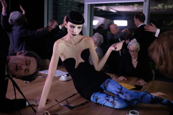 Model in a black strapless dress and blue patterned leggings lying across a wooden conference table surrounded by people in a corporate office