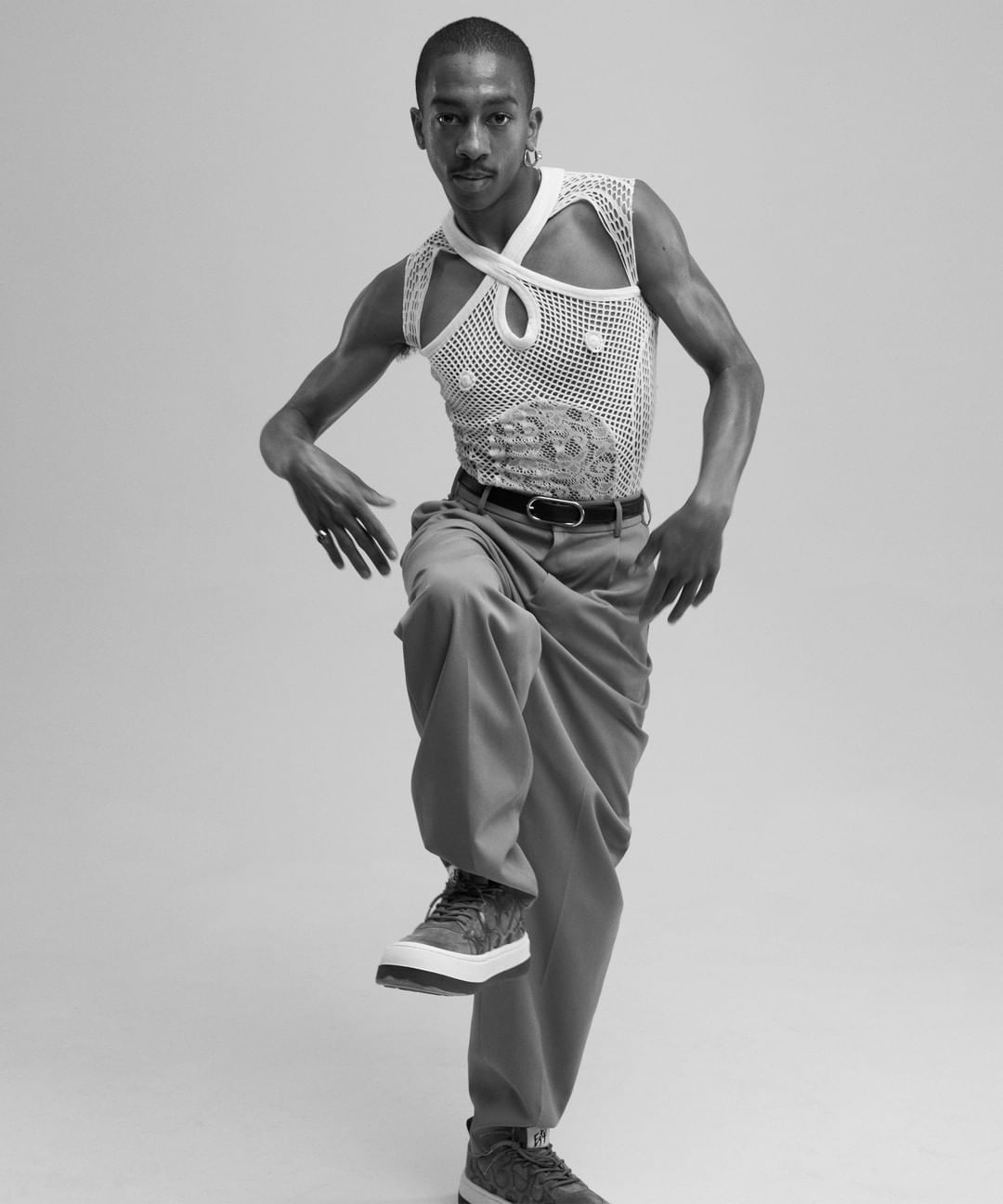 Black and white studio shot of a slender man posing in a textured white top, grey slacks and sneakers, balanced on one leg.