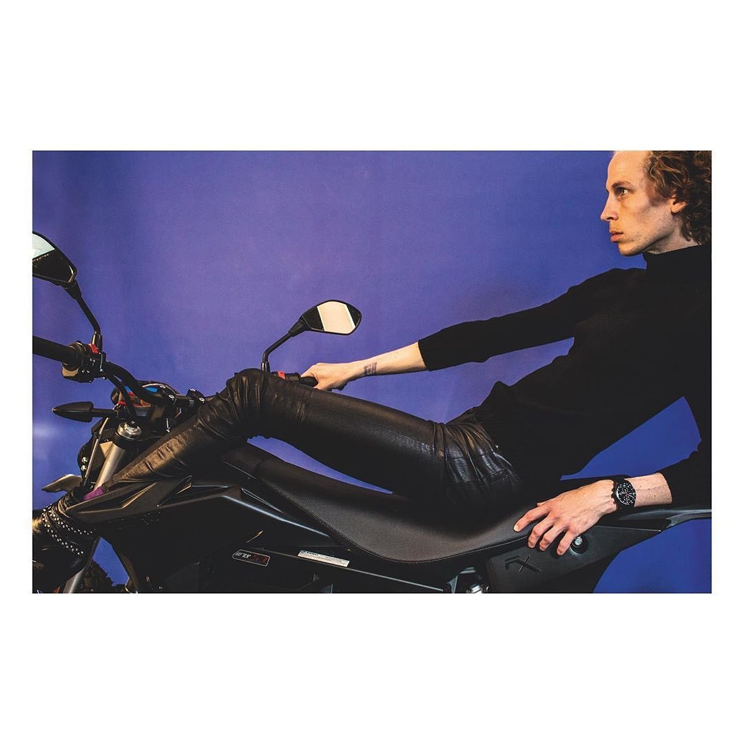 A fair-skinned male model with curly hair poses on a black motorcycle against a blue backdrop, wearing black leather pants and a black turtleneck.