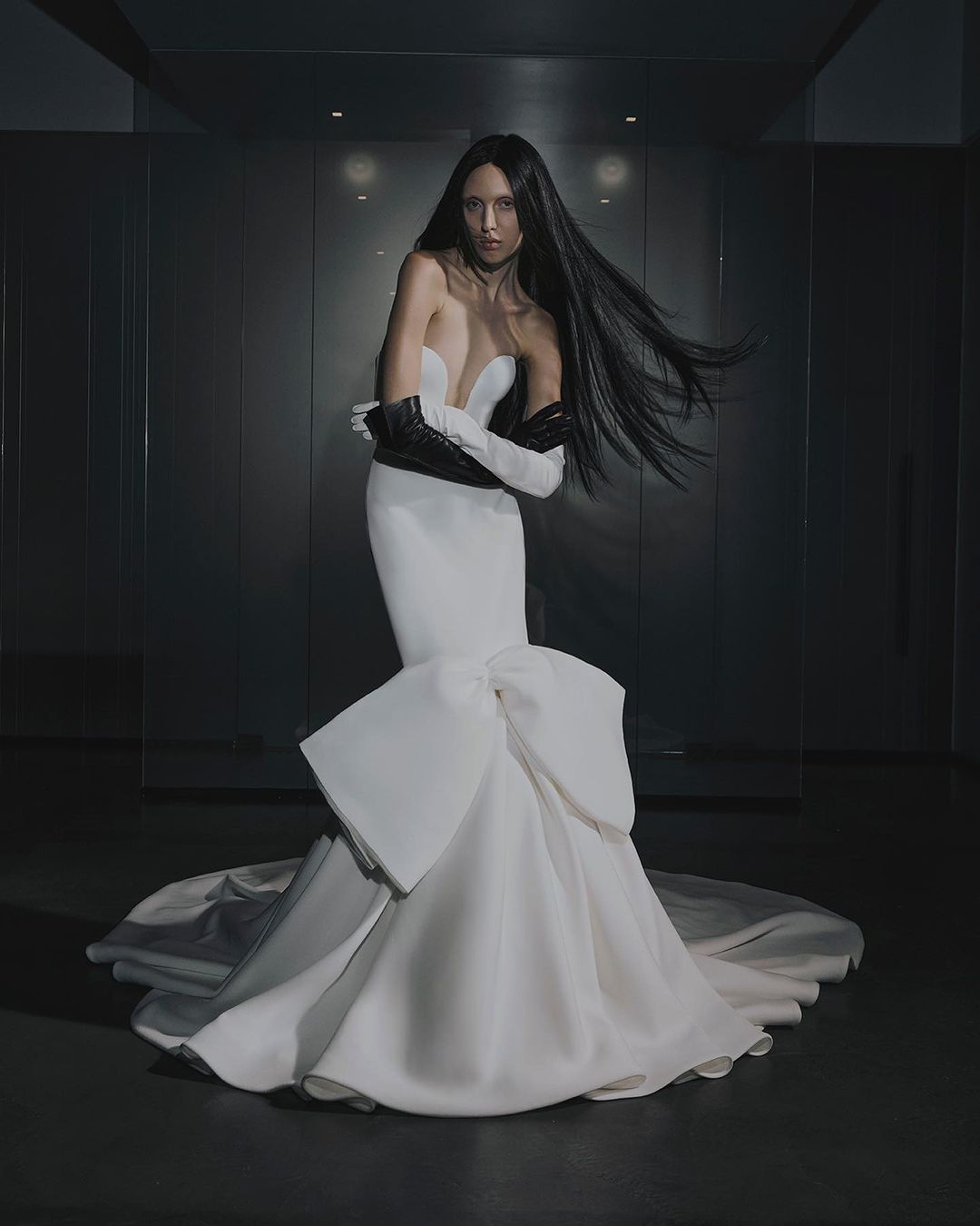 A woman with long dark hair poses in a white strapless gown with a large bow detail, wearing black opera gloves in a studio setting.