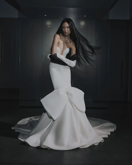 A woman with long dark hair poses in a white strapless gown with a large bow detail, wearing black opera gloves in a studio setting.
