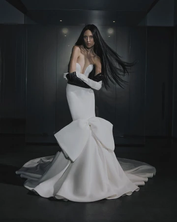 A woman with long dark hair poses in a white strapless gown with a large bow detail, wearing black opera gloves in a studio setting.