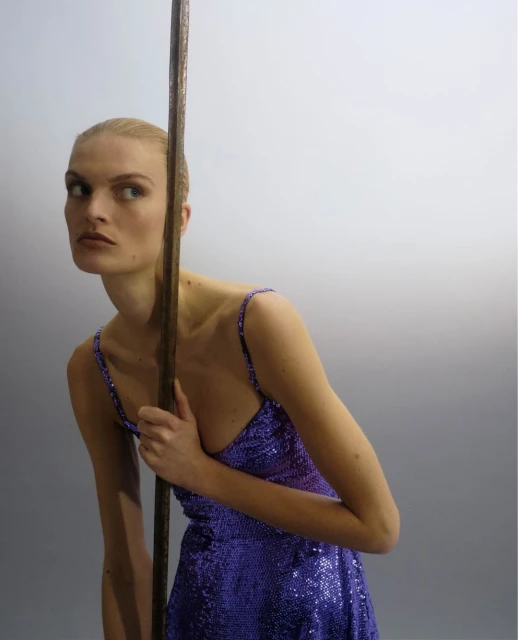 Woman in a purple sequin mini dress poses next to a tall thin, brown metal object against a white wall in a studio. Beauty shot.