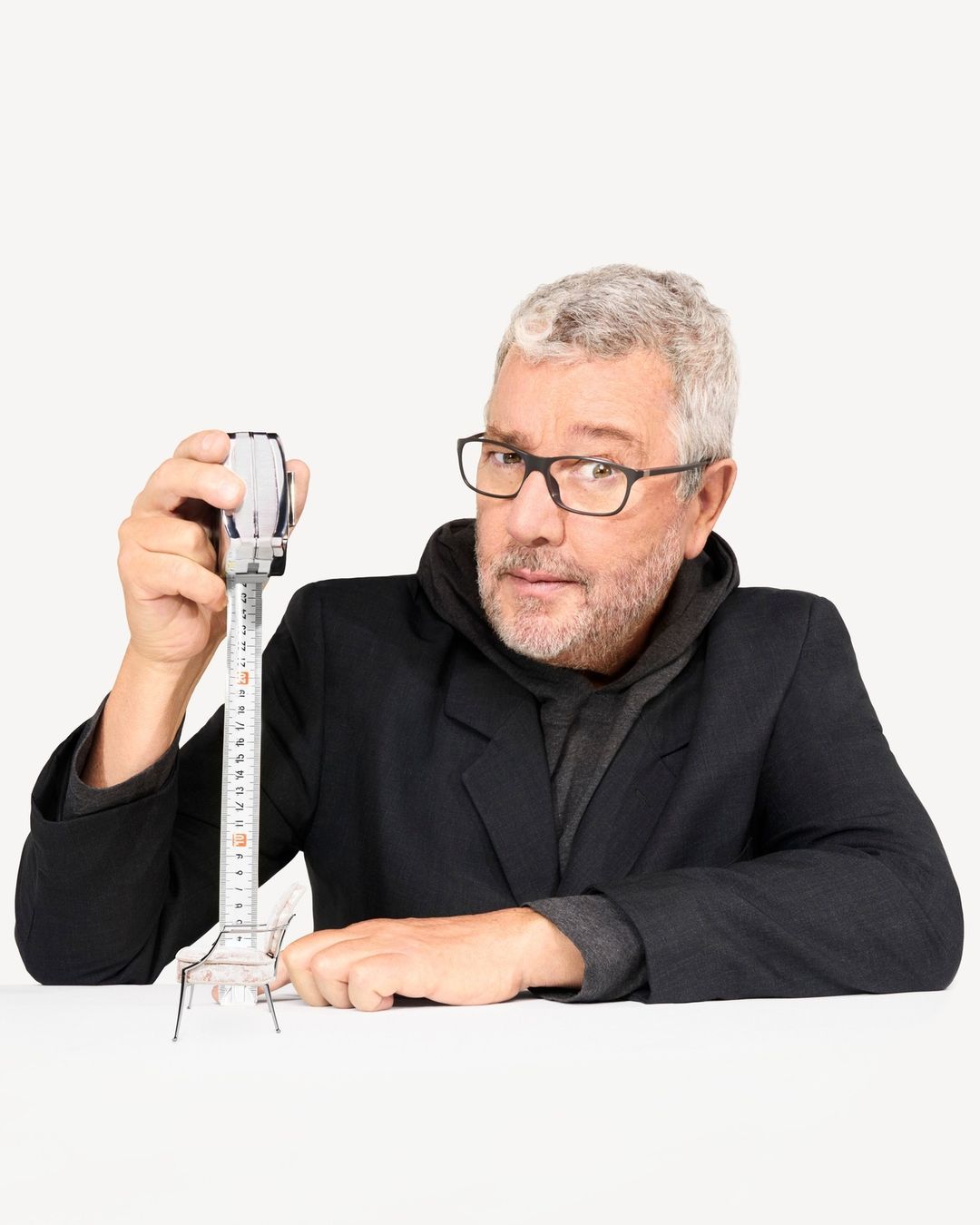 A middle-aged man with glasses and gray hair holds a measuring tape over a miniature chair against a white backdrop in a studio shot.