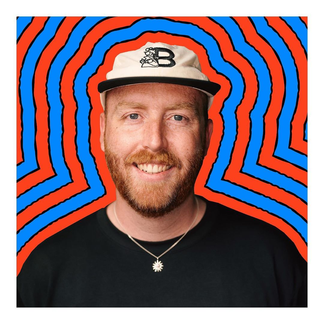 Portrait of smiling adult male with fair skin and red beard wearing a hat with a 'B' logo against a geometric red and blue background.