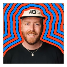 Portrait of a man with red hair and a beard wearing a white cap and black t-shirt against a vibrant blue and red illustrated background.