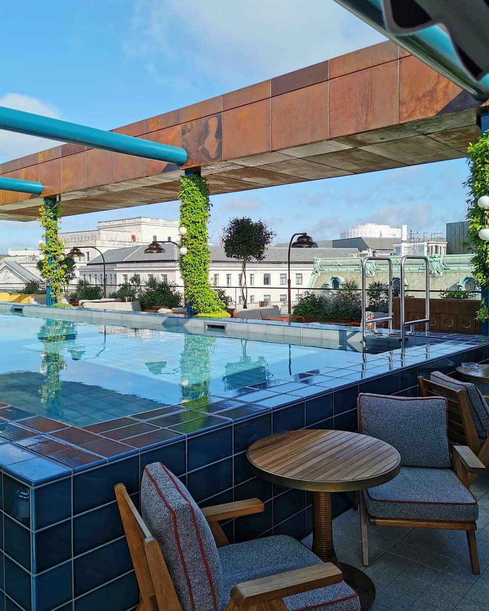 Rooftop swimming pool with blue tiles and lounge chairs on a sunny day. Ivy covered pillars and city buildings are visible in the background.