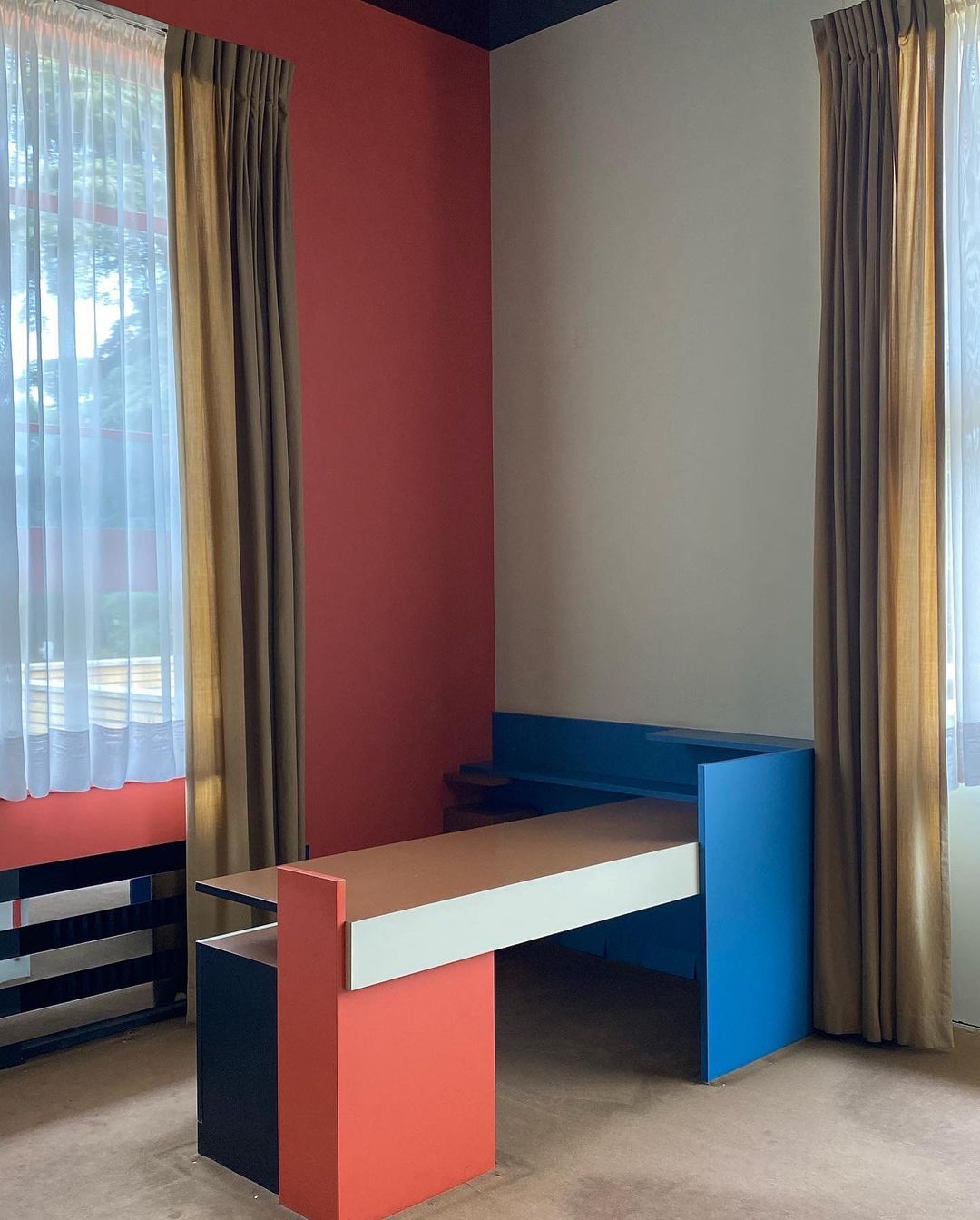 Colorful geometric interior featuring a modern desk with orange, white, and blue accents, set against a red and beige wall, with curtains.