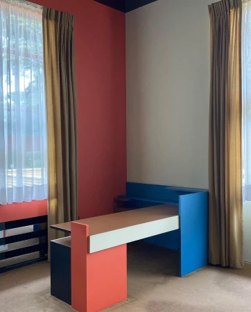 Colorful geometric interior featuring a modern desk with orange, white, and blue accents, set against a red and beige wall, with curtains.