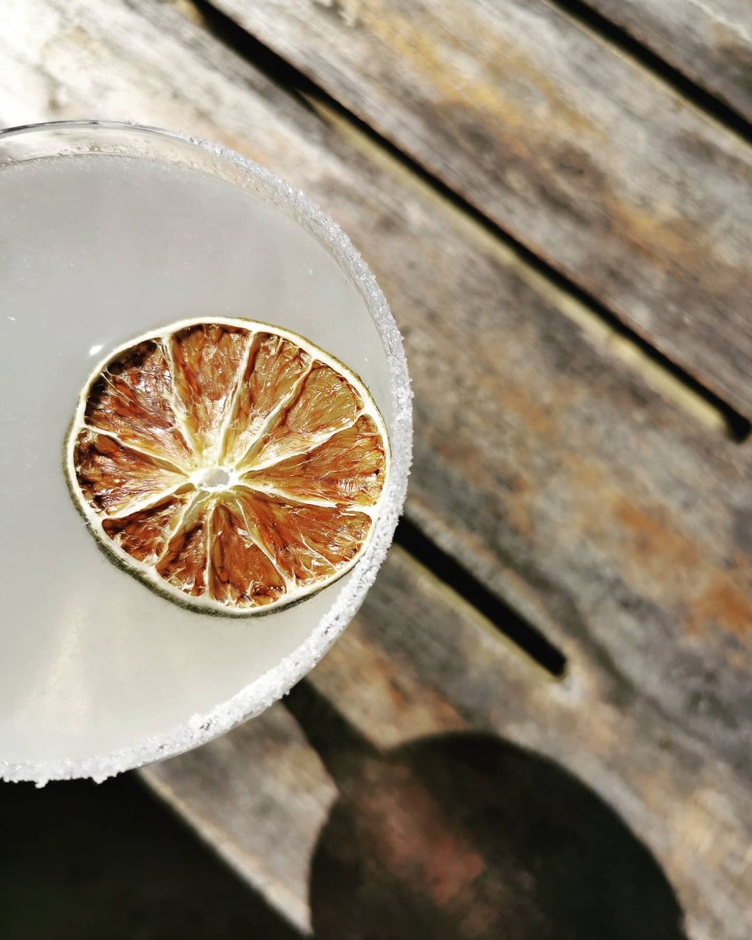 Overhead shot of a margarita in a glass with salted rim and dried lime wheel garnish on rustic wooden planks in natural light.