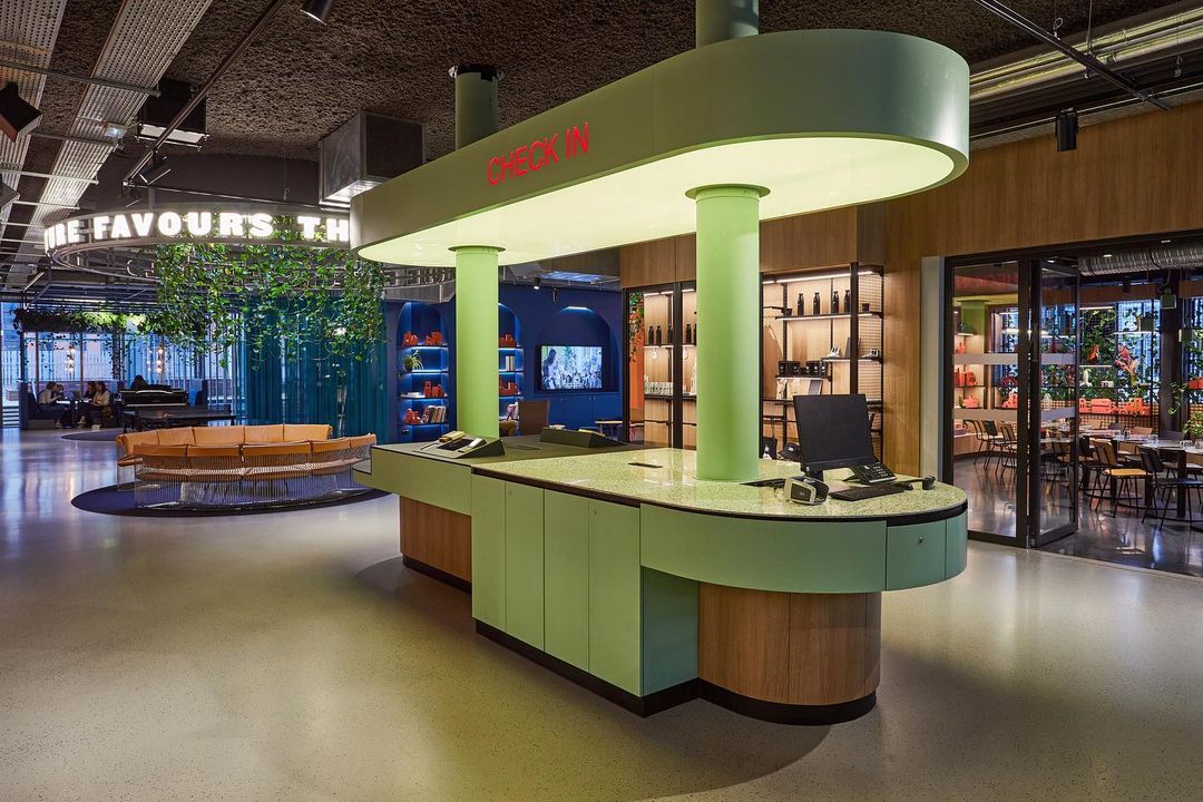 Interior view of a hotel lobby featuring a light green check-in desk, decorative greenery, and stylish seating in a modern design.