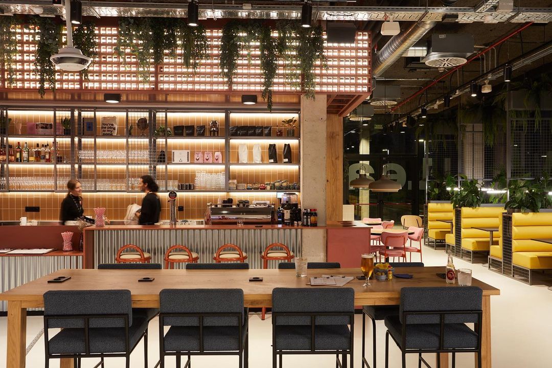 Modern indoor cafe and bar with two people behind the counter, tables and chairs with plants hanging from the ceiling.