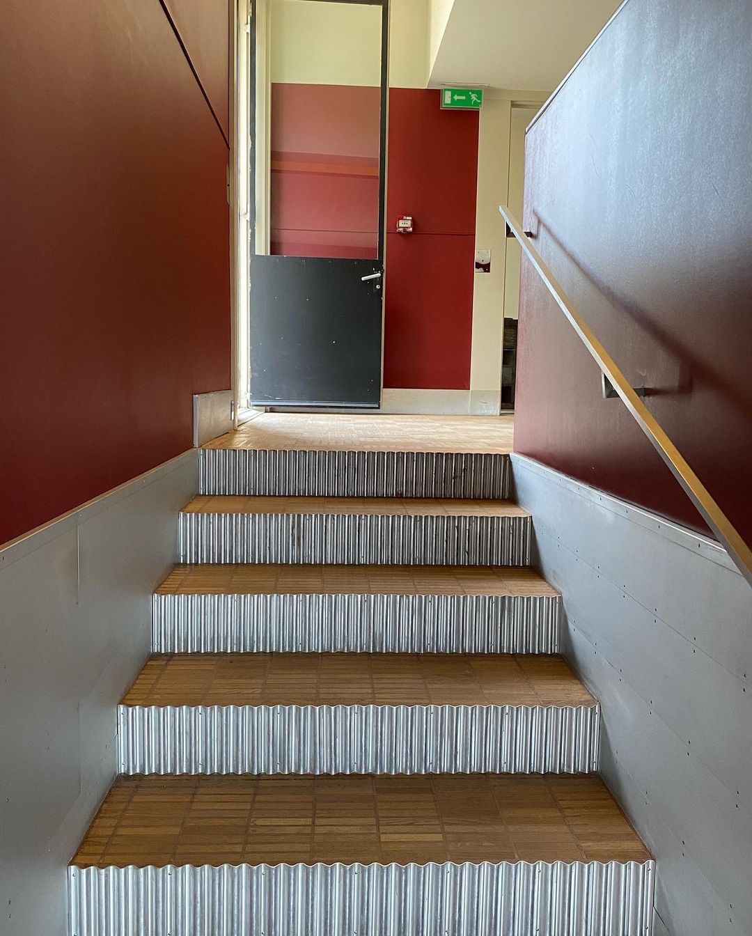 Indoor shot of stairs with corrugated metal risers and wooden treads leading to a landing with maroon walls and a door.