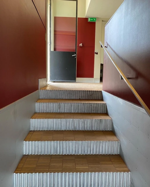 Indoor shot of stairs with corrugated metal risers and wooden treads leading to a landing with maroon walls and a door.