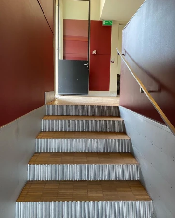Indoor shot of stairs with corrugated metal risers and wooden treads leading to a landing with maroon walls and a door.