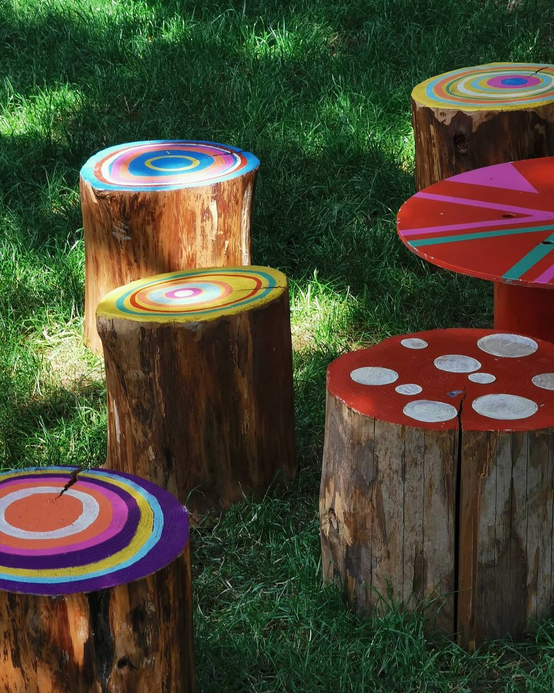 Colorful, painted tree stumps arranged outdoors on a lawn. Natural wood textures contrast with bright geometric designs.