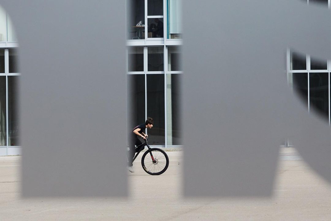 Person on a bicycle rides past a modern building with large windows, partially obscured by a large, grey, stencil-cut letter.