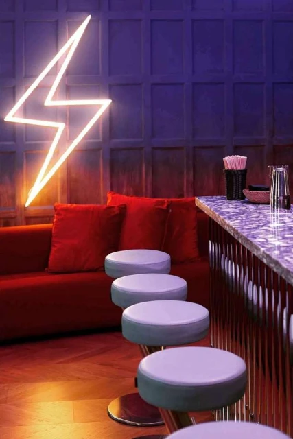 Bar scene featuring red sofa, round stools, marble counter, and neon lightning bolt sign against a textured blue paneled wall.