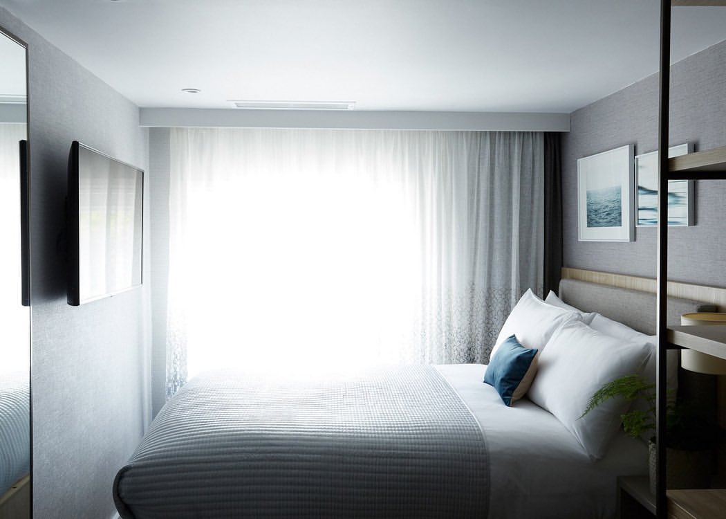 Bedroom interior featuring a queen-sized bed with white linens, blue throw pillow, a flat-screen TV, and soft natural light through sheer curtains.