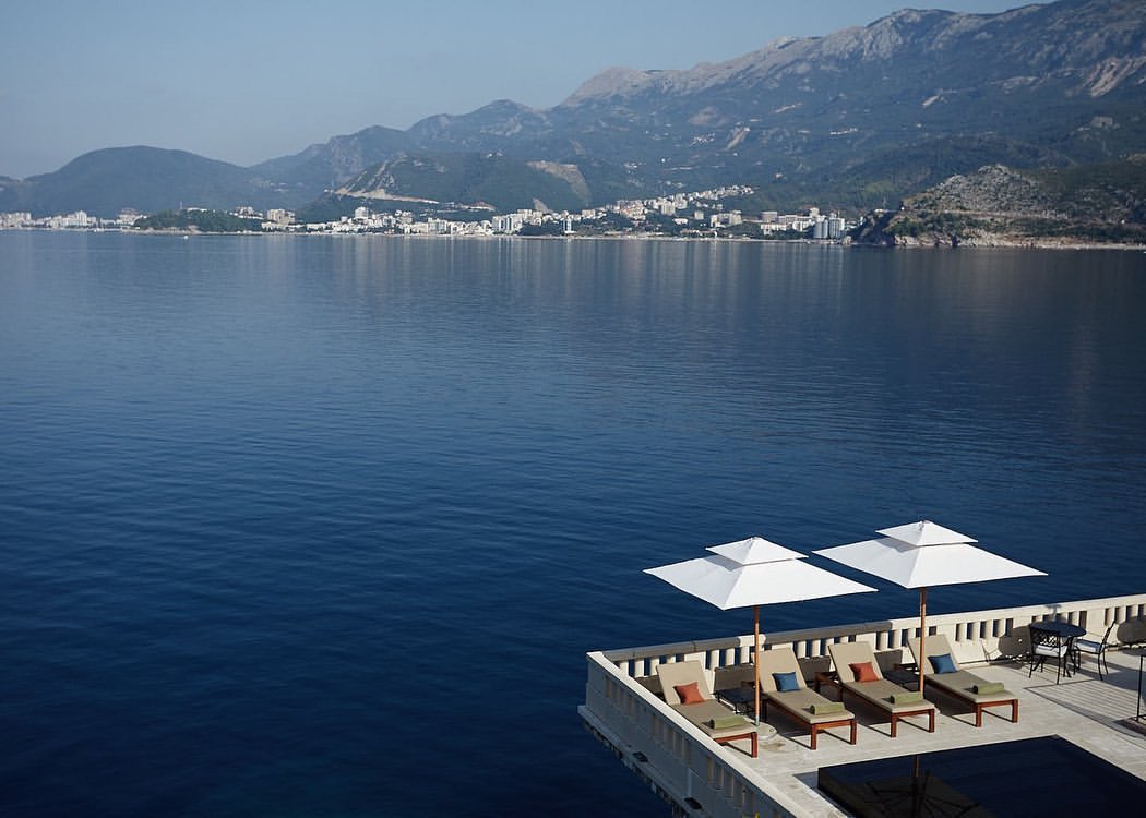Seascape view of tranquil blue water, mountains, and a coastal town, featuring lounge chairs with white umbrellas on a deck.