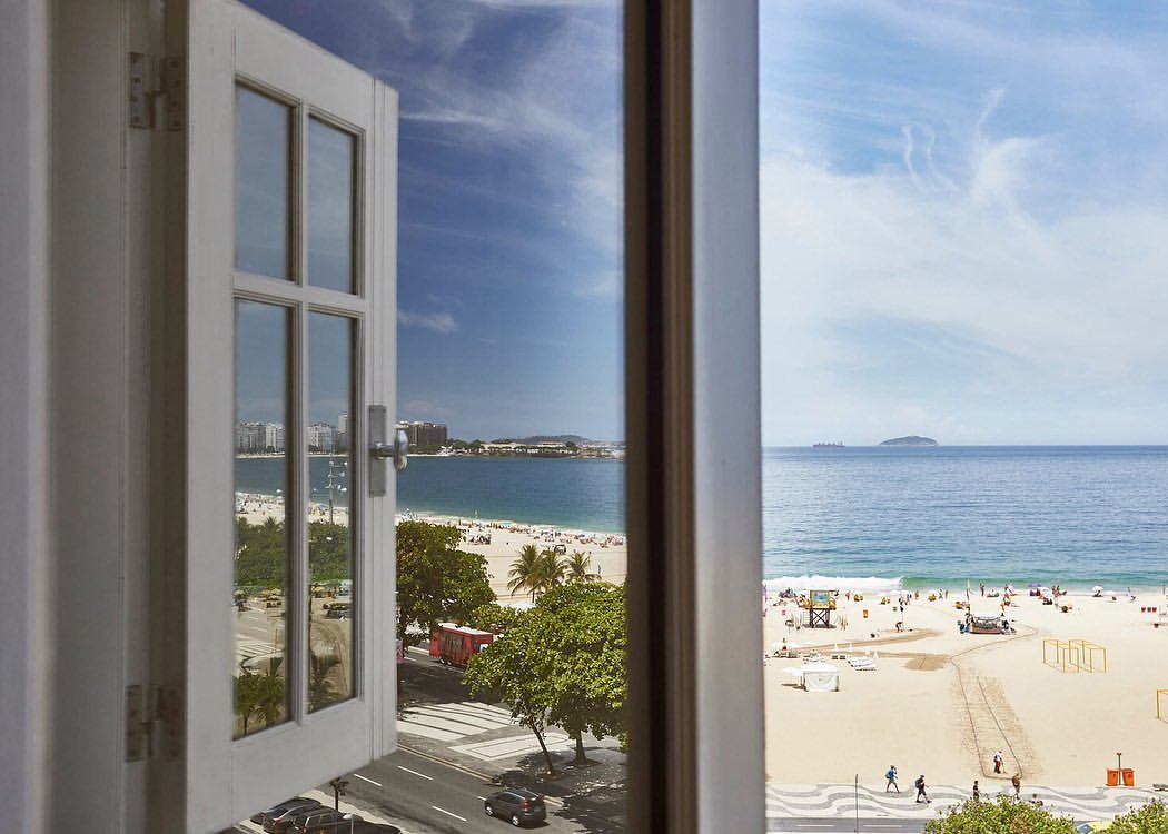 View from open window of a tropical beach with sand, people, and water; city skyline in the distance, blue sky with clouds.