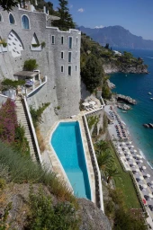 High-angle view of a stone cliffside hotel featuring a long swimming pool overlooking the blue ocean water and coast.