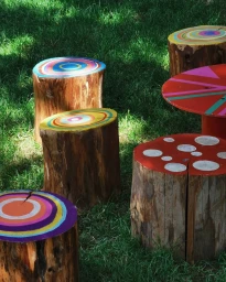 Painted tree stump stools and a table arranged in green grass outdoors in London by Tim Evan-Cook.