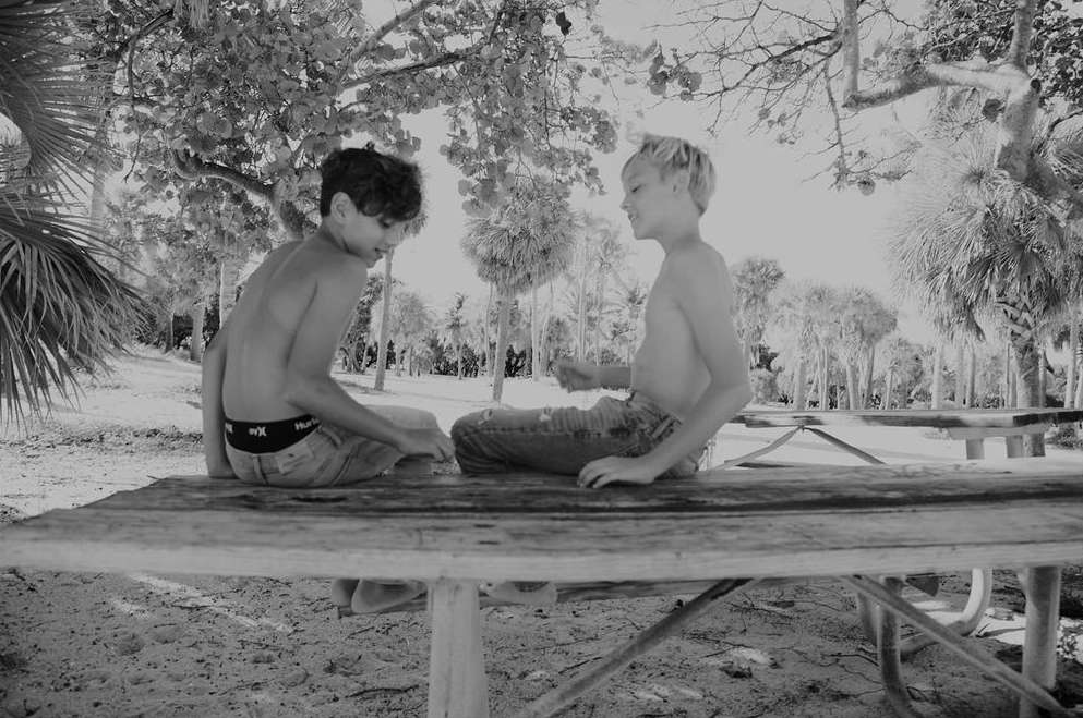 Two shirtless boys sit on a wooden picnic table outdoors surrounded by palm trees. The monochrome image has a shallow depth of field.