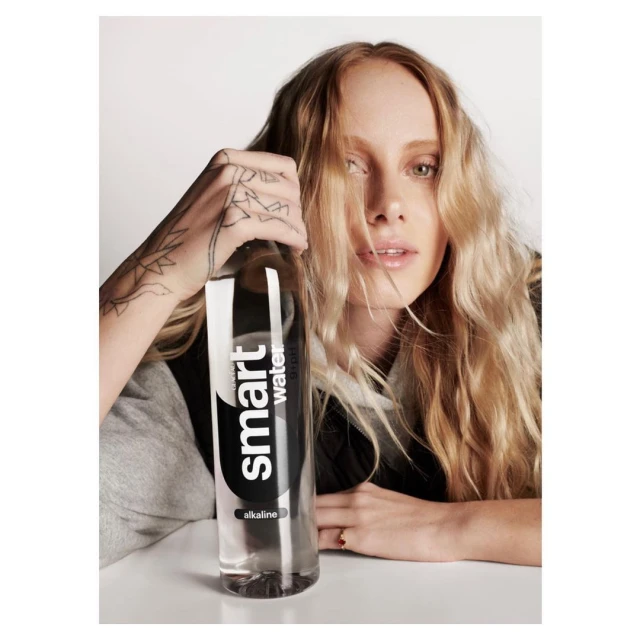 Woman with blond hair poses indoors with Smartwater bottle, wearing a grey sweatshirt, against a solid white backdrop.